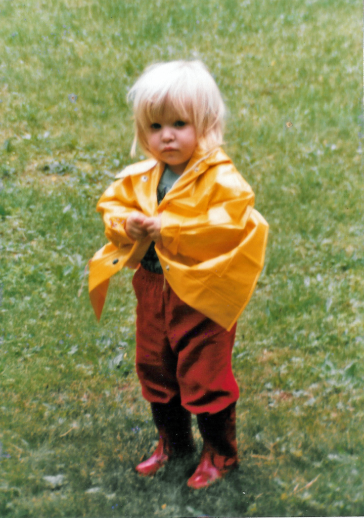 a small white, blonde child, maybe around 2 years old, wearing red rain boots, red pants, and a yellow rain jacket