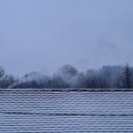 a dusting of snow on a roof with chimney smoke and trees in the background