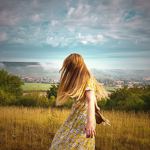 a white lady with long blonde hair blowing over her face while she twirls, behind her is grass and hills and a light blue sky with wispy white clouds