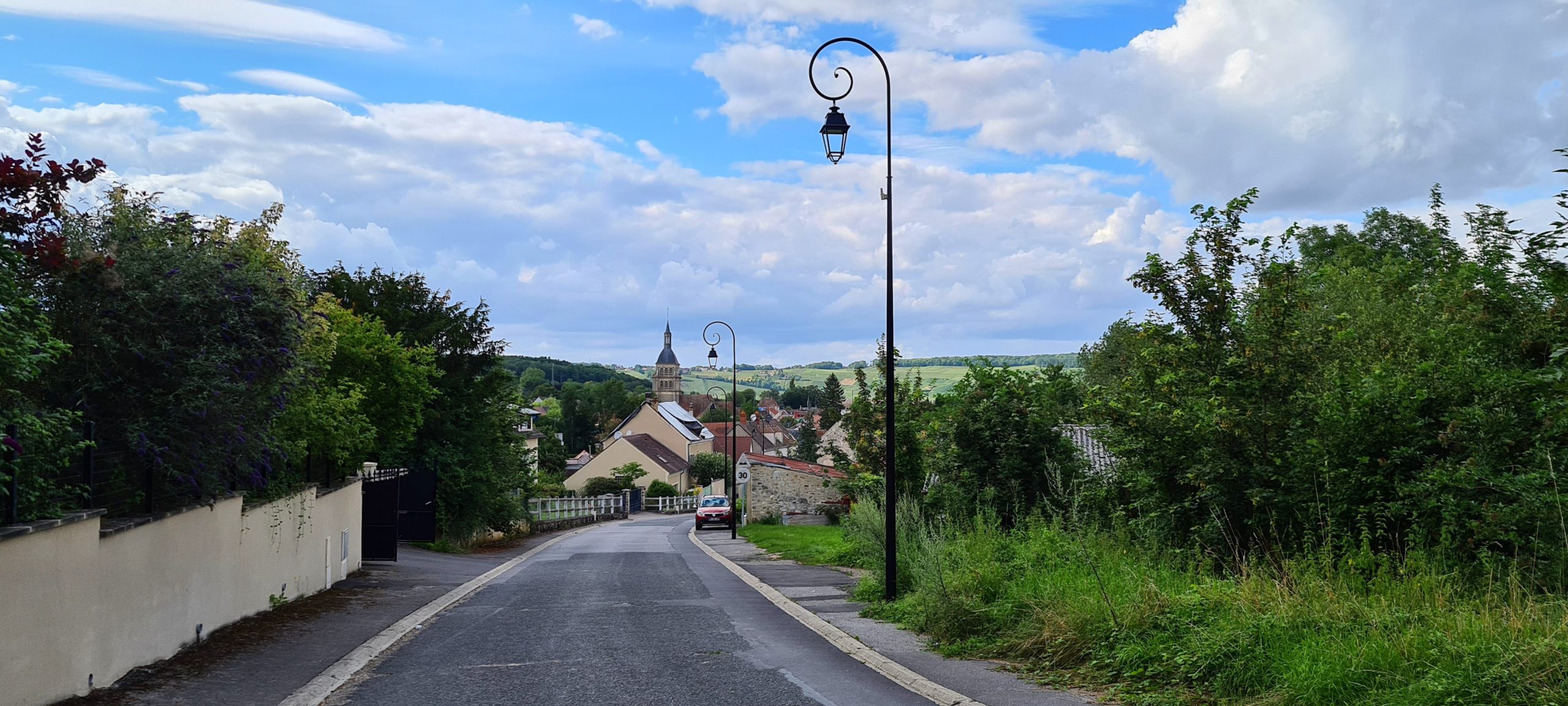 a road to a small village with a church steeple in the background and decorative street lights along the way