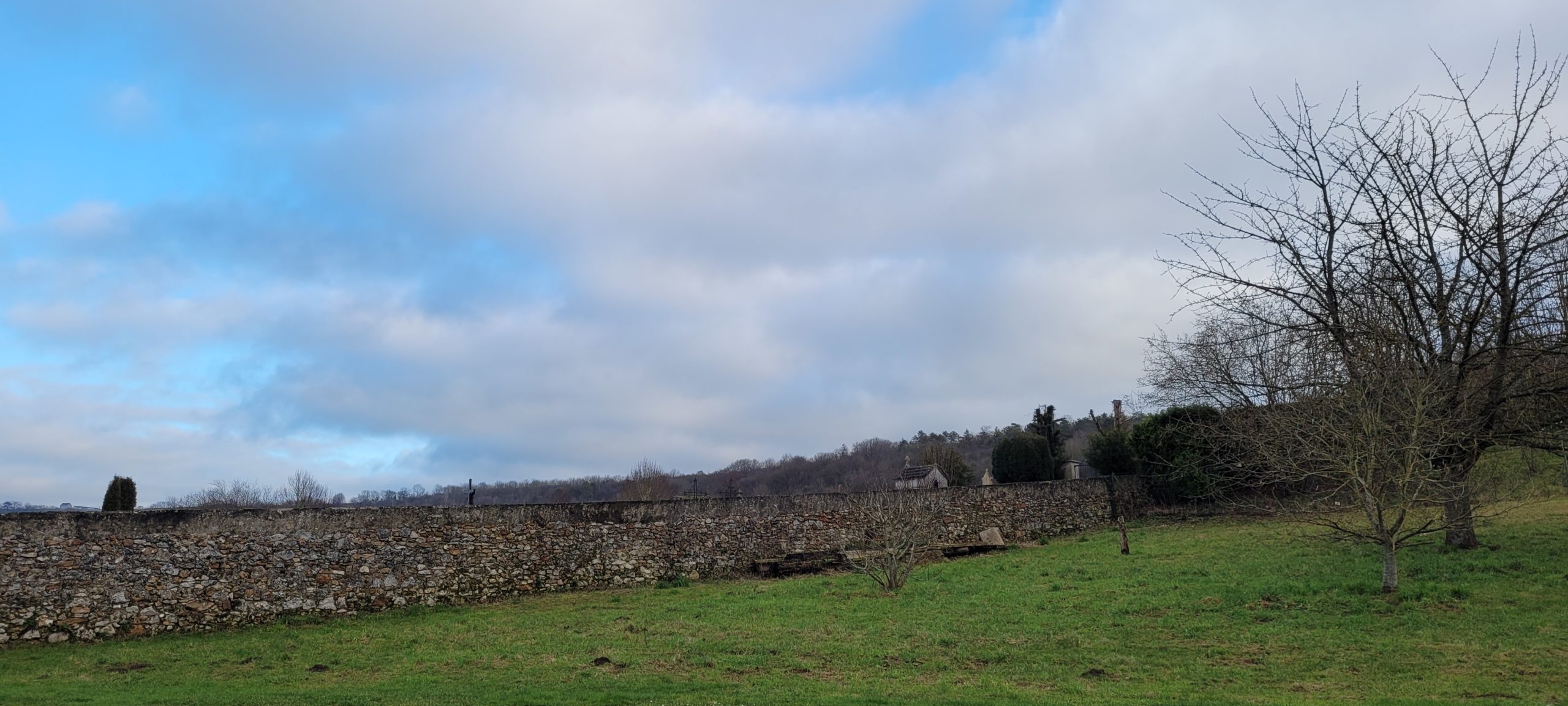 blue sky, white clouds, green grass and rock wall hiding a cemetery