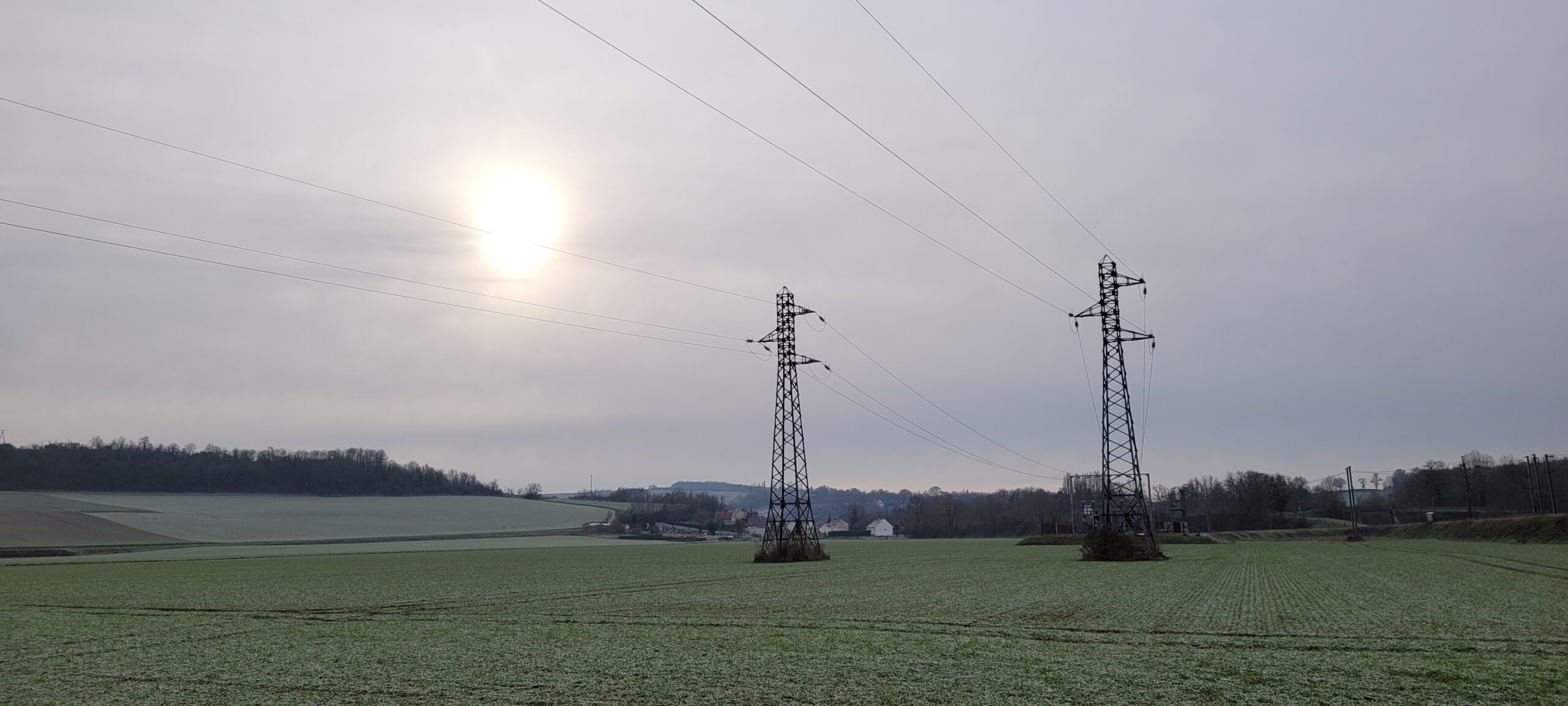 power lines/towers in farm land in front of a cloudy sky obscuring the sun